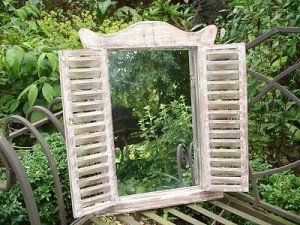 A shabby chic mirror with shutters sitting on a bench in a garden.