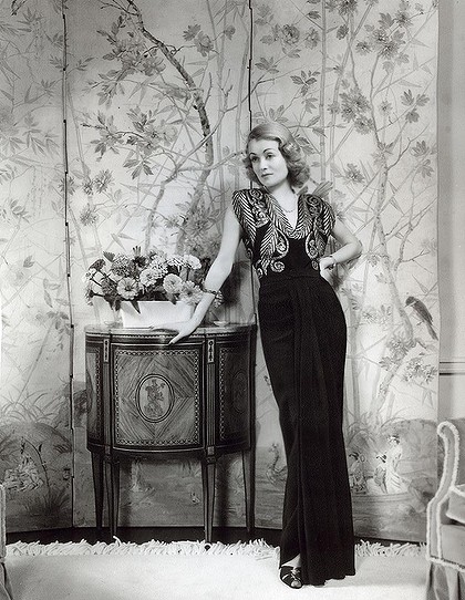A woman in a black dress posing in front of a hollywood regency wallpaper.