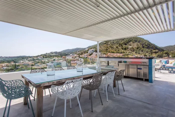 On this rooftop terrace by Fantastic Frank, a sleek outdoor kitchen features stainless steel appliances and a built-in barbecue. It faces away from the lounge area, while a pergola offers dining shade and modern chairs in white, gray, and blue enhance the contemporary vibe.