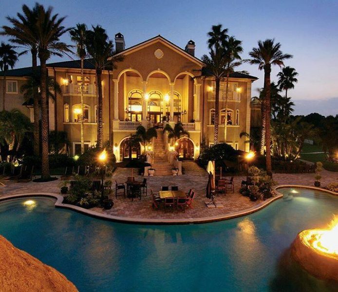 A luxurious mansion at dusk with a pool and fire pit.