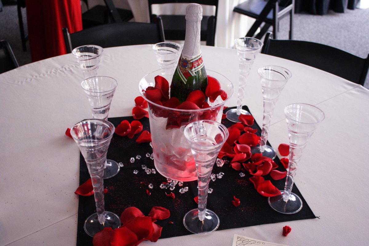 A table adorned with Valentine's Day decor features a bottle of champagne surrounded by red rose petals.