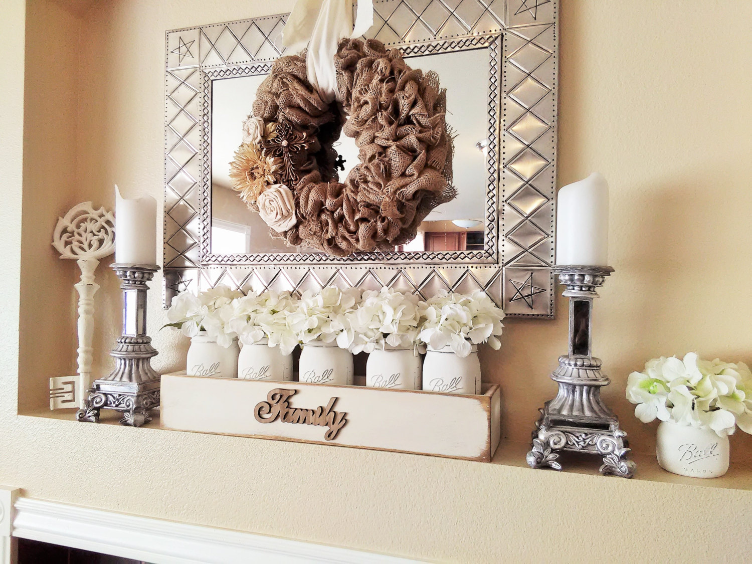 A mantel decorated with a burlap wreath and mason jars filled with flowers.