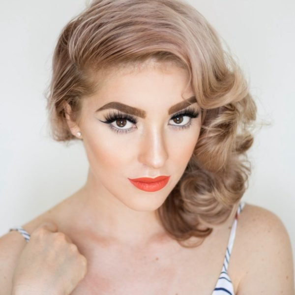A woman with blond hair and orange lipstick posing for a photo, showcasing her eyelash extensions.