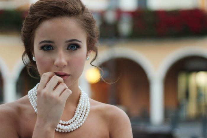 A young woman wearing a white dress and pearls adds a touch of blush to her natural beauty.
