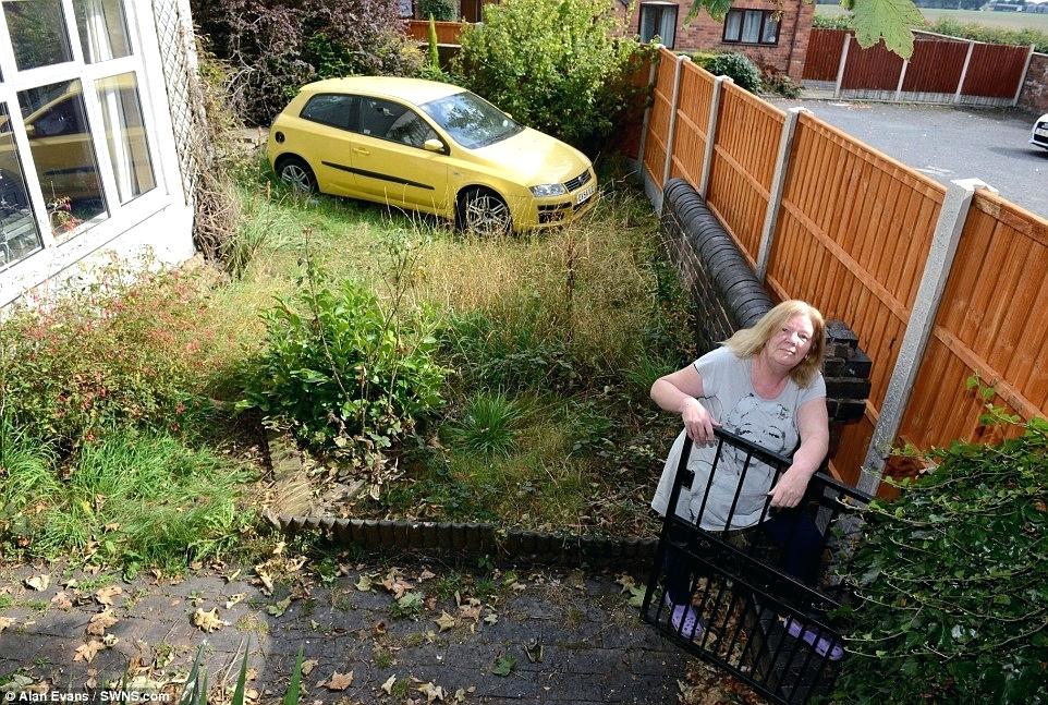 A woman standing next to a yellow car on a property line fence.