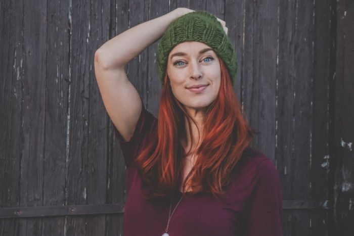 A woman with red hair and dark circles wearing a green knitted beanie.