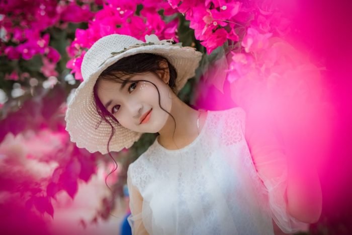 A girl in a white hat posing in front of pink flowers and drinking water.