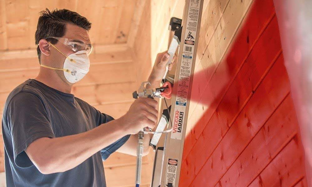 A man, wearing a mask, is using a paint sprayer to paint a red wall.