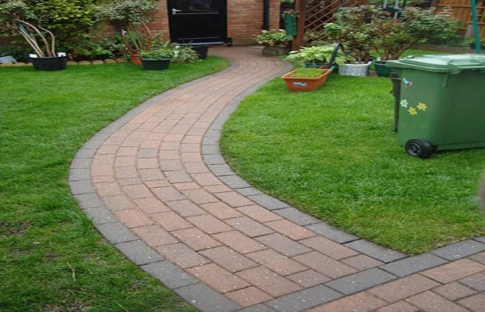 A garden with a brick path and a green trash can, paving.
