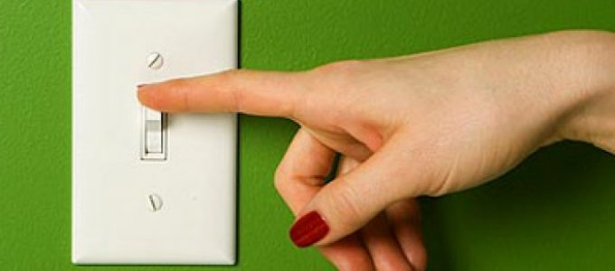 A hand is pointing at a light switch on a green wall, encouraging to save electricity.