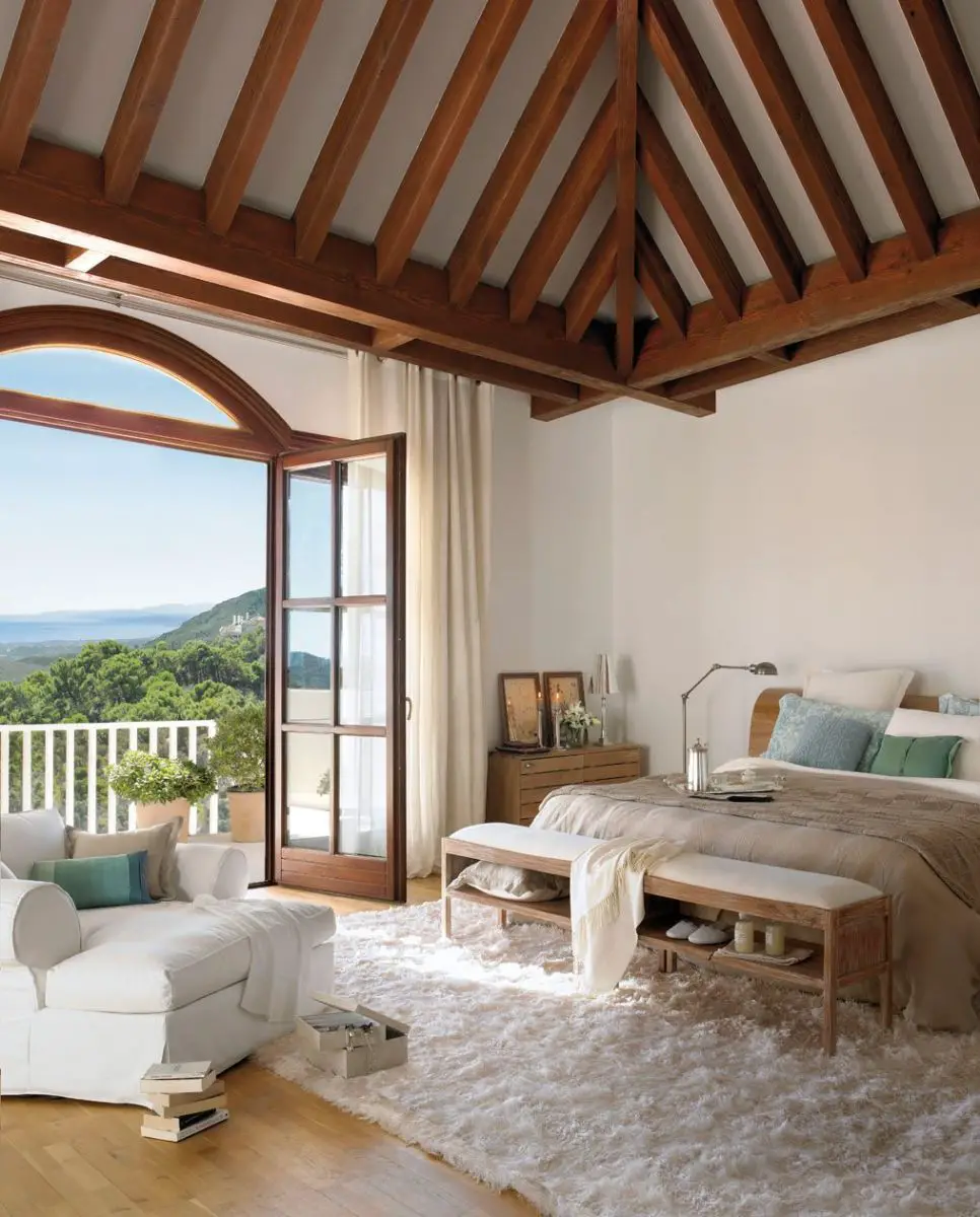 A white bedroom with wooden beams and abundant natural light from the ocean view.