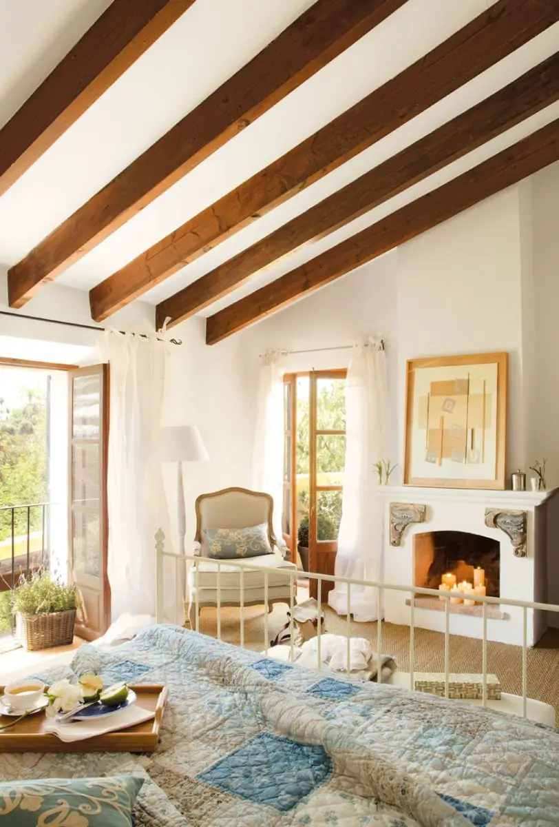 A bedroom with natural light, wooden beams, and a fireplace.