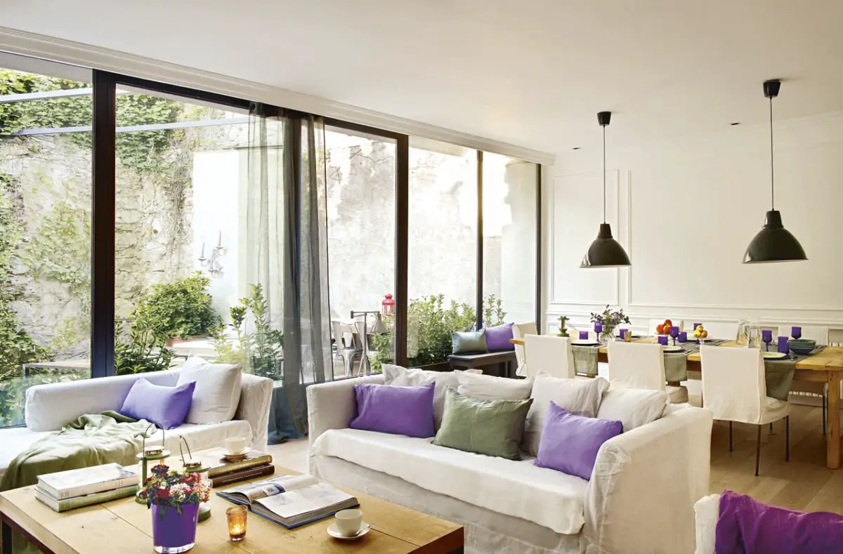 A living room with white furniture and purple pillows, adorned with curtains.
