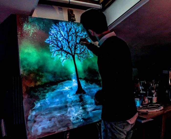 A man painting a tree in a dark room using unique paints.