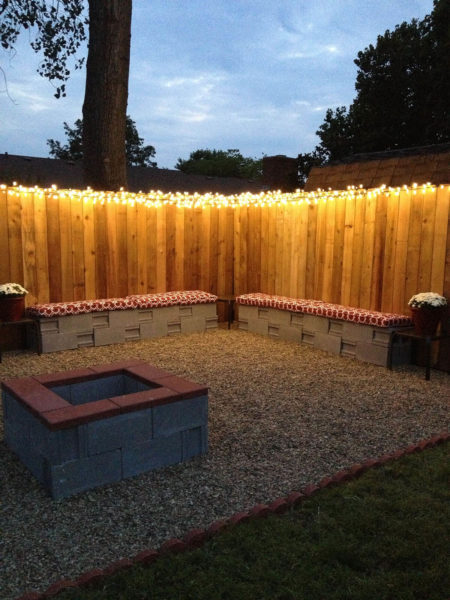 A backyard with a fire pit and benches illuminated by outdoor lighting.