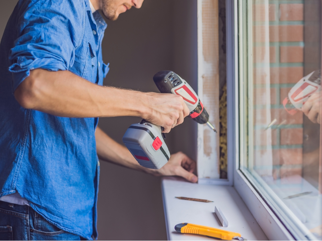 A man performing repairs at home using a drill on a window sill.