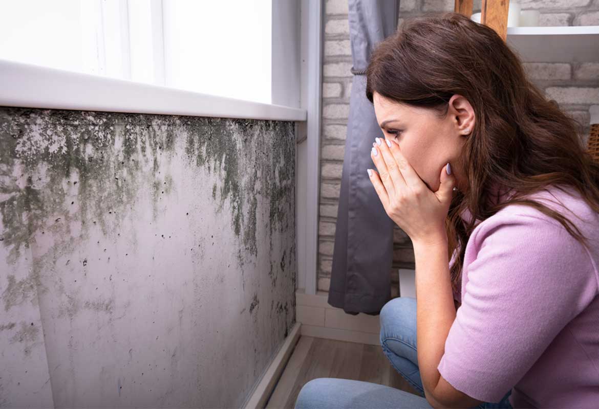 A woman sneezing in front of a moldy wall.