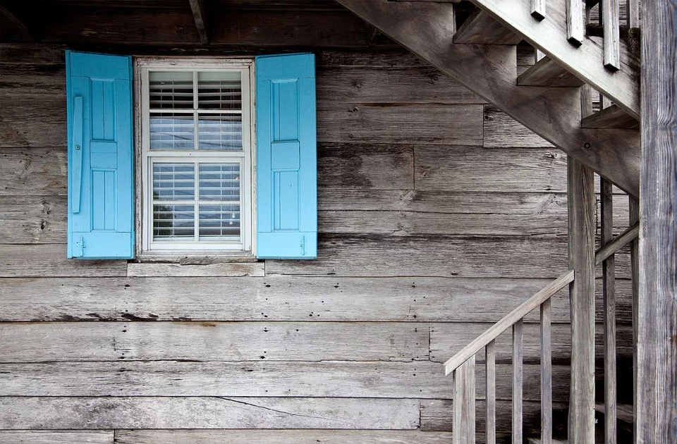 How custom home builders can help you realize your dream house by incorporating a blue shuttered window on a wooden house.