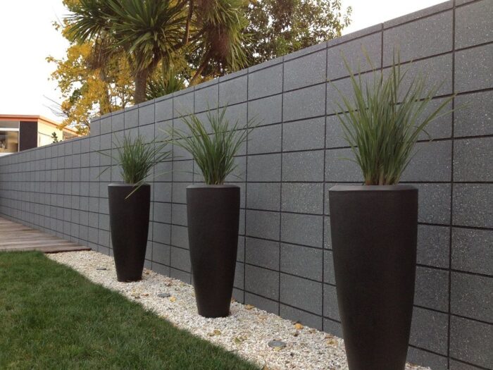 Three black potted plants in front of a gray wall with architectural design.