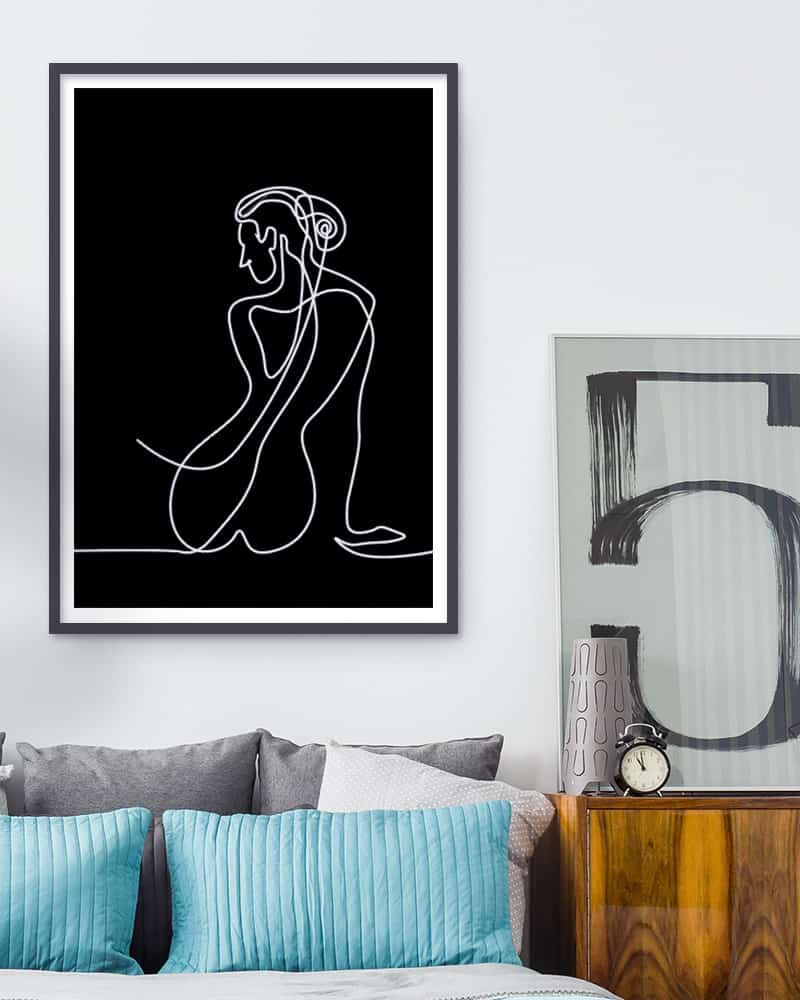 A black and white drawing of a woman sitting on a bed, with pictures on wall.