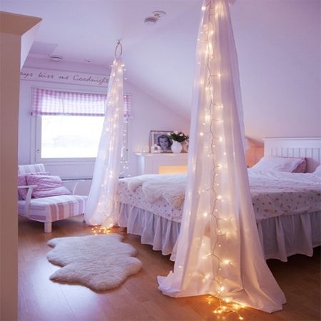A bedroom with string lights illuminating a bed covered in white curtains.
