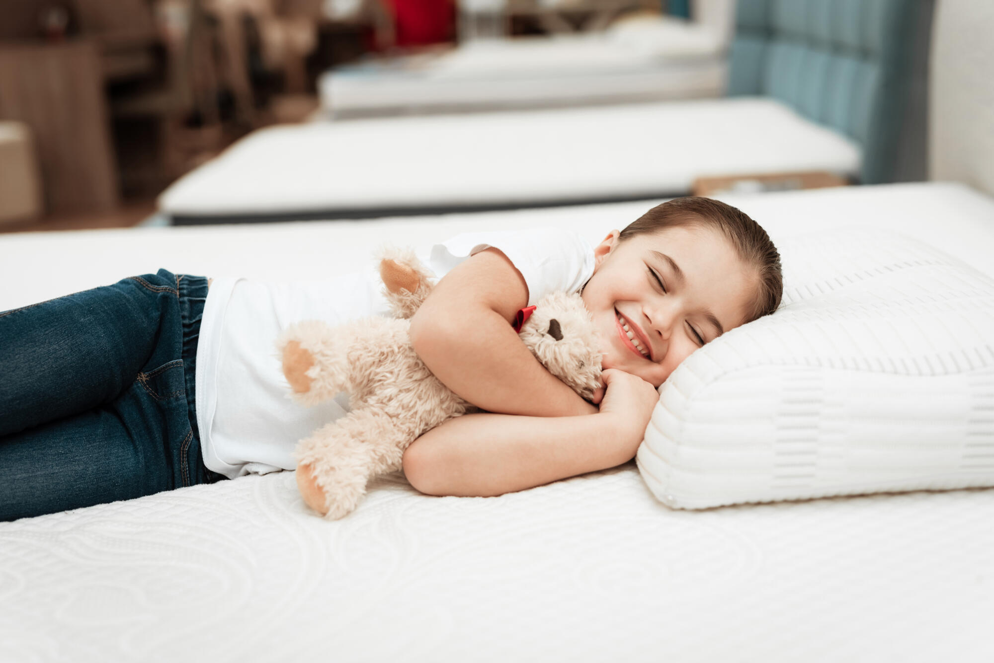 Smiling little girl lies on n orthopedic mattress in a furniture store. Smiling little girl hugs teddy bear on mattress in furniture store.