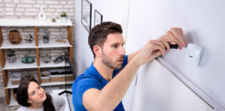 Happy Woman Looking At Electrician Installing Security System Motion Detector On Wall