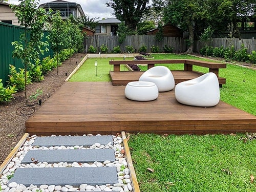 A backyard with a wooden deck and chairs, featuring landscaping.