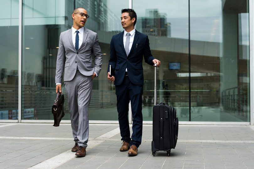 Two businessmen on a business trip walking with suitcases in front of a building.