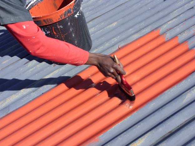 A man is painting an orange roof with a brush using anti-corrosive paint to protect it from rusting.