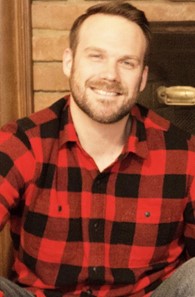 A man sitting in front of a fireplace wearing a red plaid shirt.