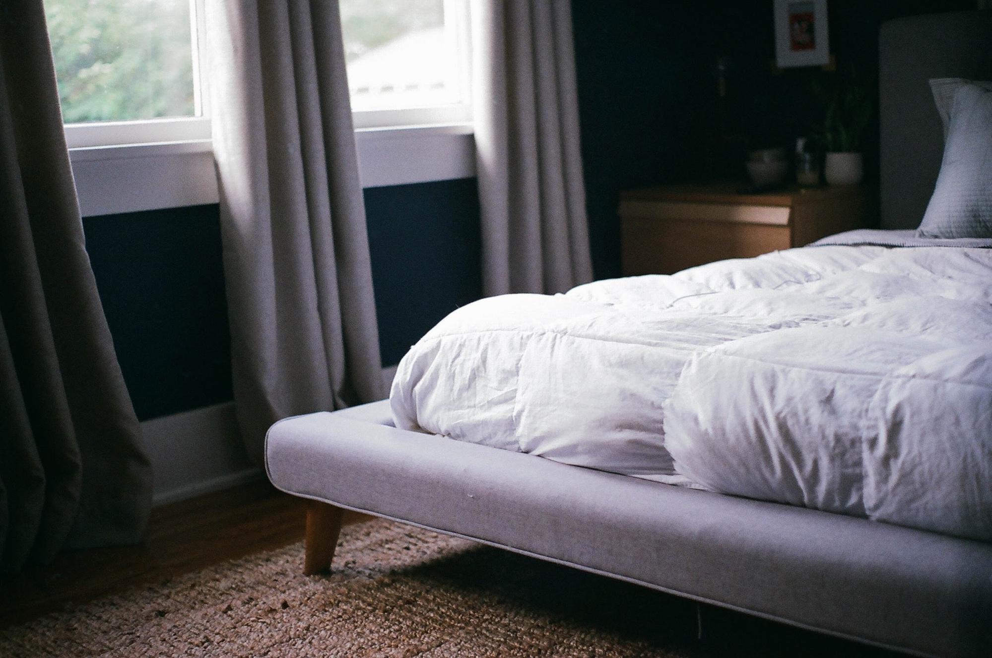 A comfortable white comforter on a bed for a good night sleep next to a window.