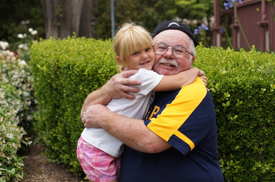 An older man hugging a little girl in front of bushes.