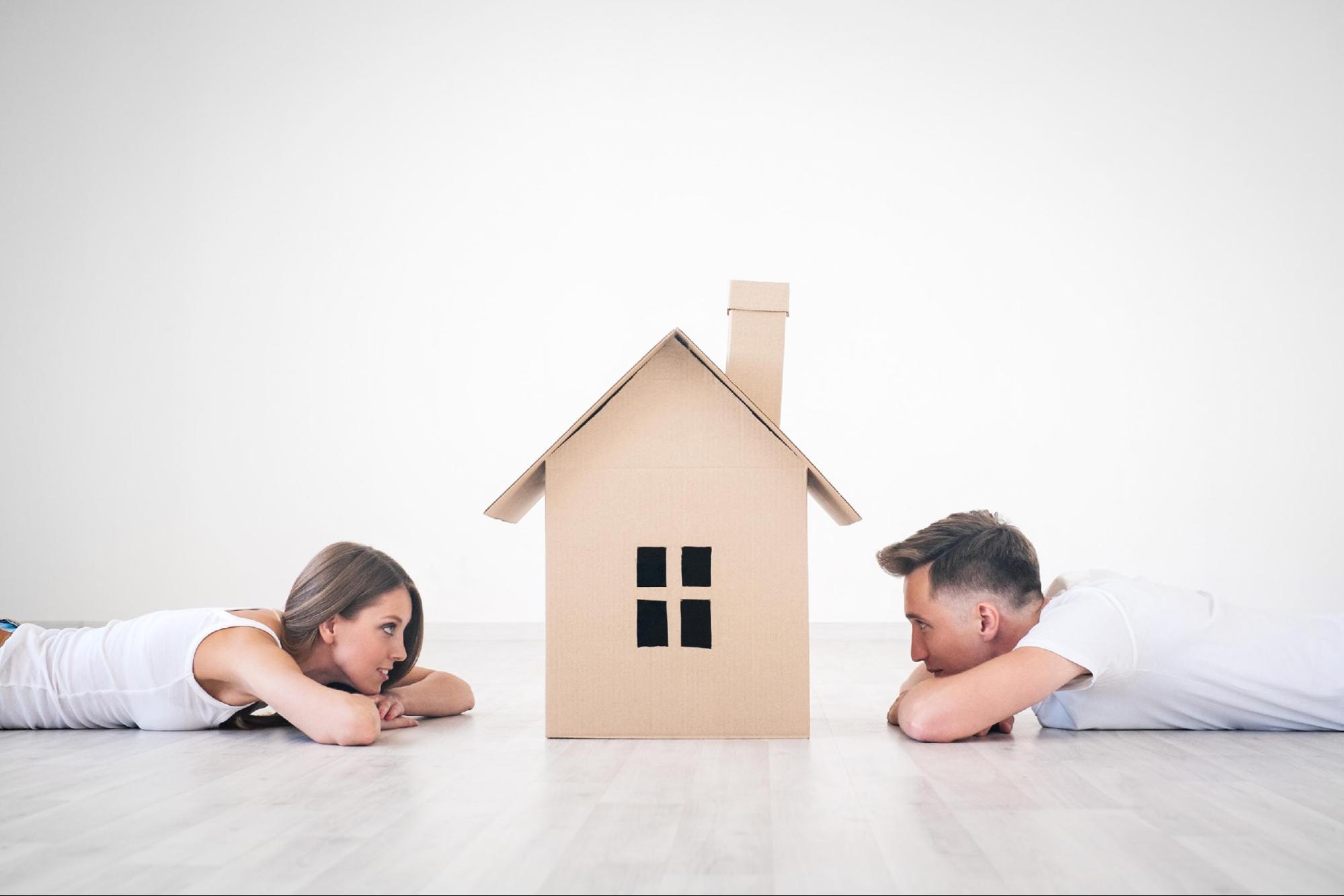Young couple laying on the floor with cardboard house in front of them, illustrating Homes For Sale In Chesapeake.