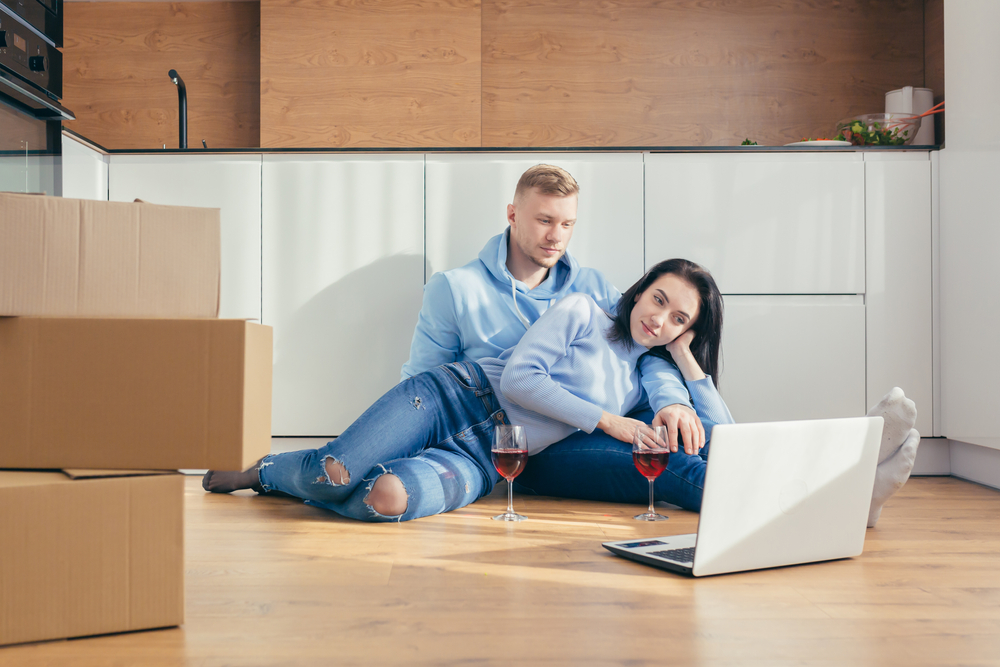 A couple Moving Into New Home sitting on the floor of their new home with a laptop.