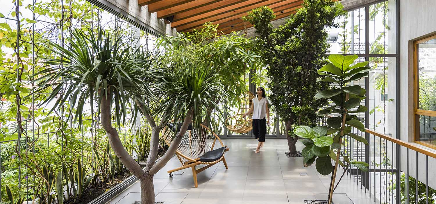 A woman is sitting on a bench in a house with a lot of plants, highlighting the importance of biophilic design.