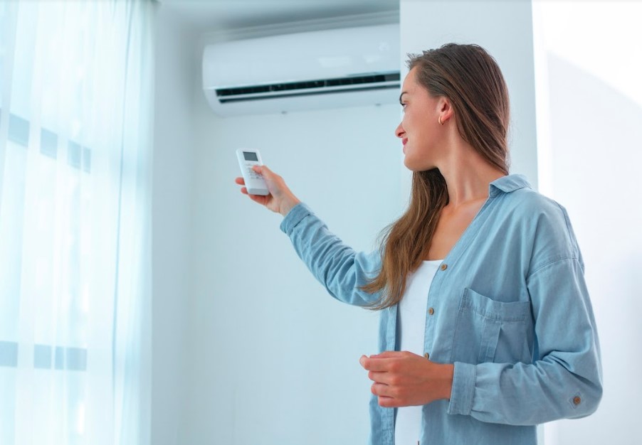 A woman holding a remote control in front of a window, preparing to adjust the air conditioner.