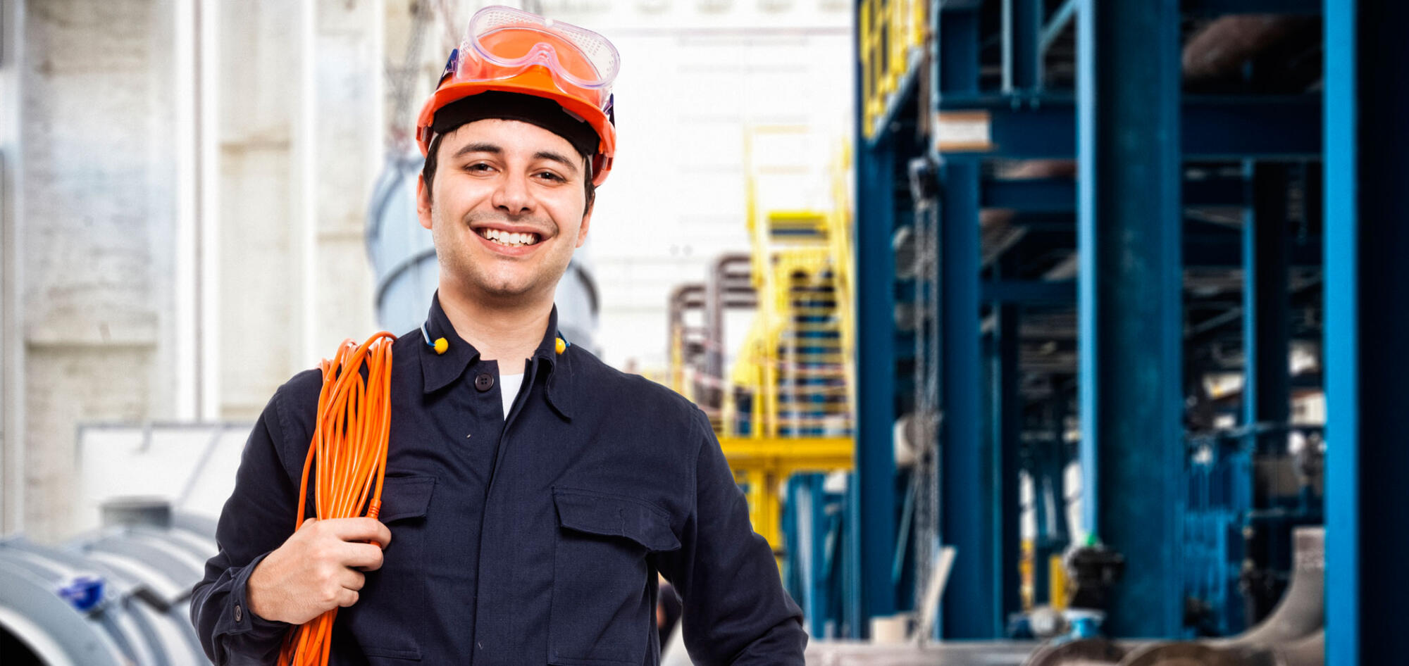 An electrician in a hard hat is smiling in front of a factory.