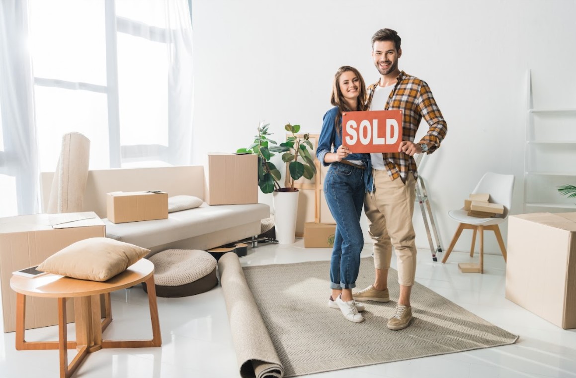 Young couple holding a sold sign in their new home; reasons people sell homes.