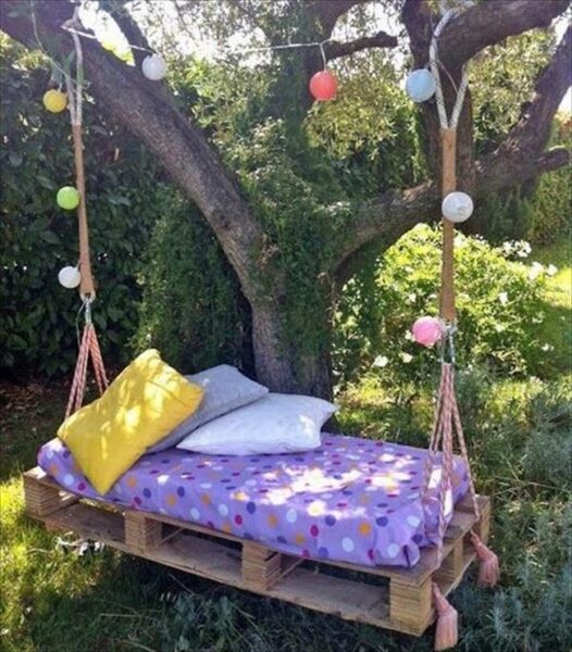 A swing bed made out of recycled pallets hanging from a tree in the garden.