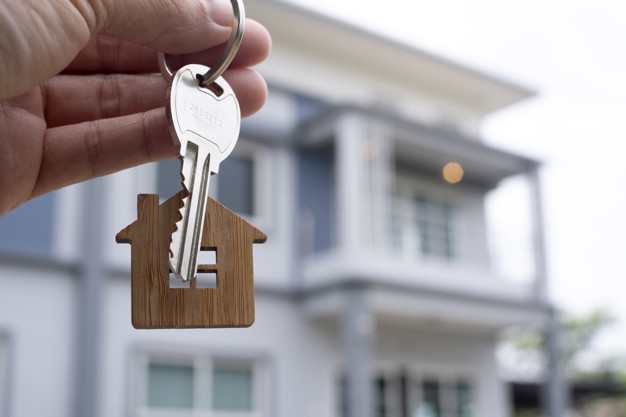 A person holding a house key in front of a house, representing the decision between renting and owning a home.