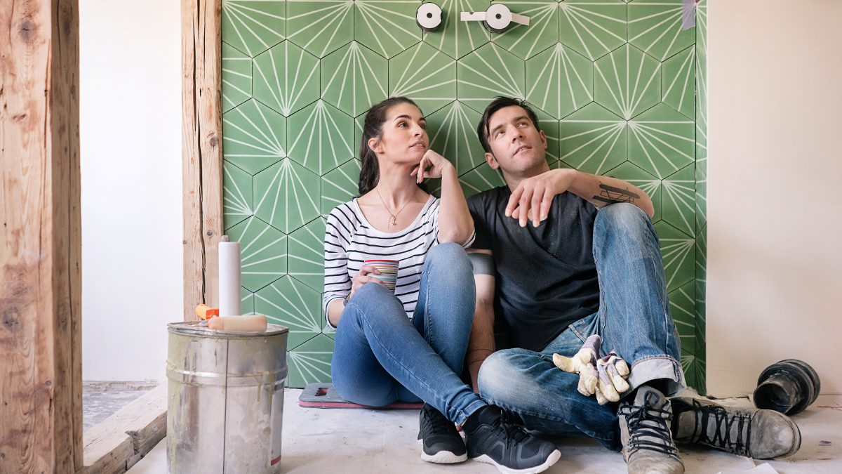A couple sits on the floor in front of a renovated green tiled wall.