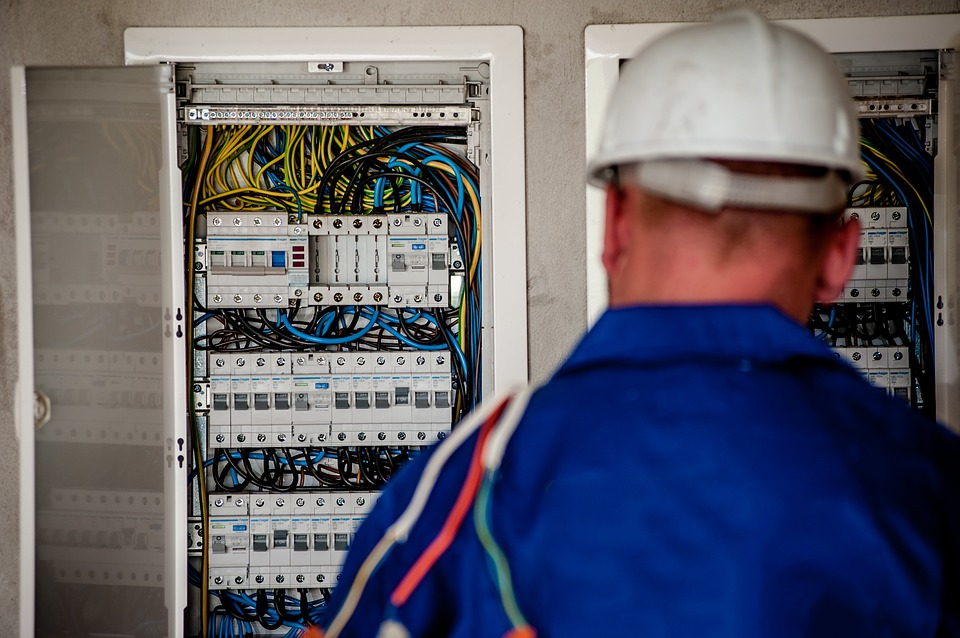 An electrician in a hard hat is standing in front of an electrical panel.