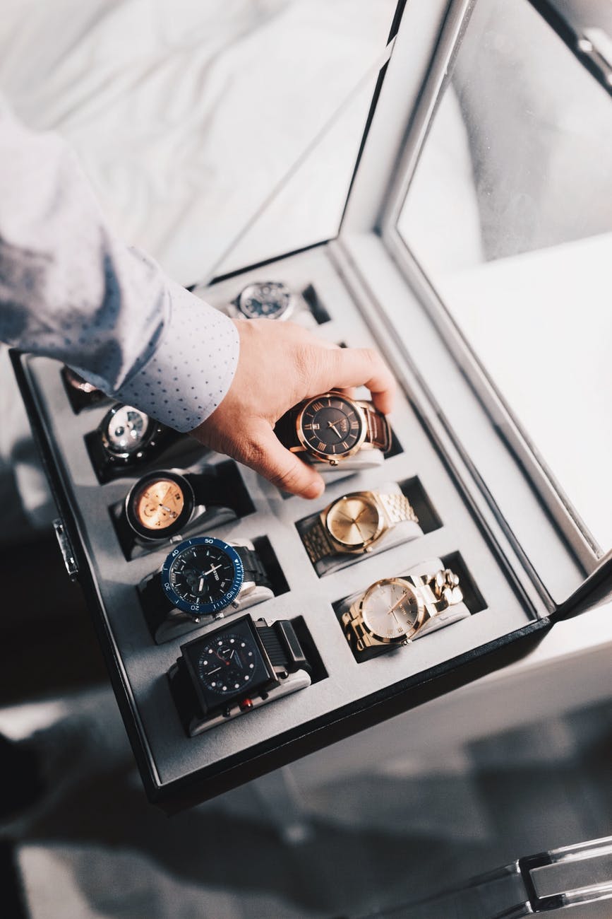 A man is placing luxury watches in a case on a bed.
