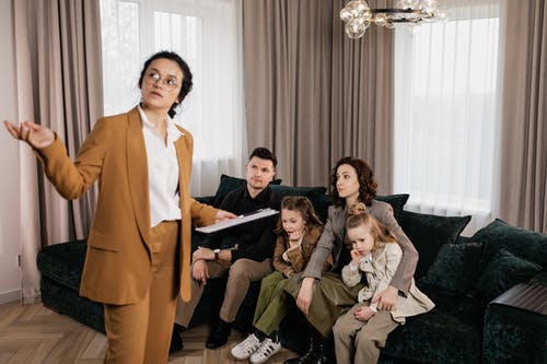 A family sits on a couch in their unsold house while a woman talks to them.