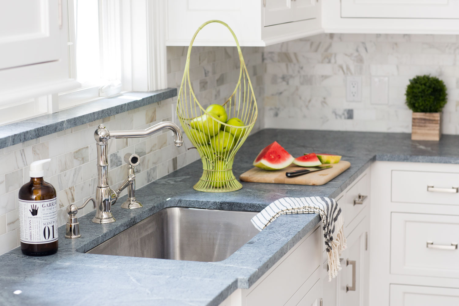 A kitchen with a sink and watermelon on the counter featuring a soapstone backsplash.