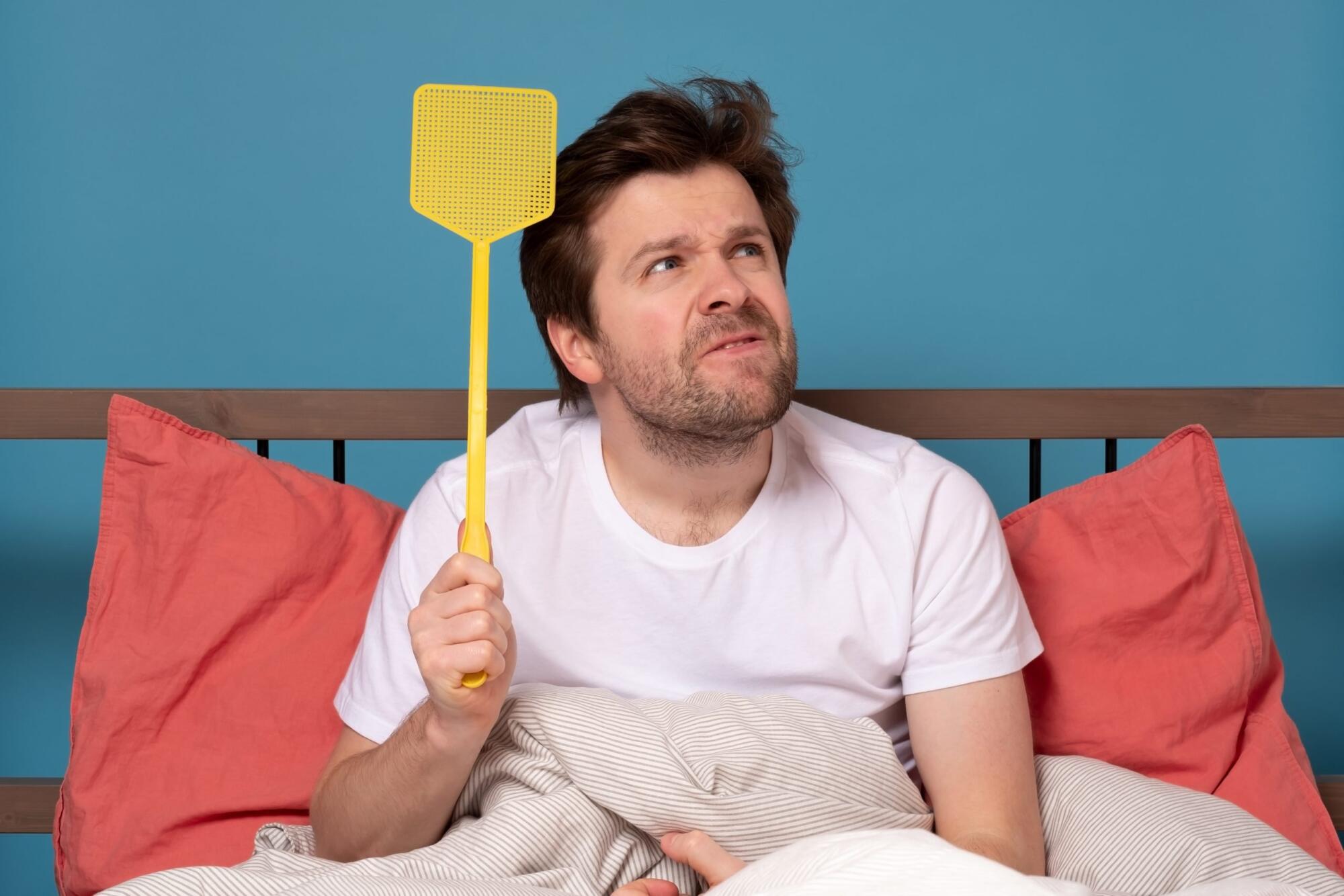 A man in bed using a yellow spatula for mosquito control.