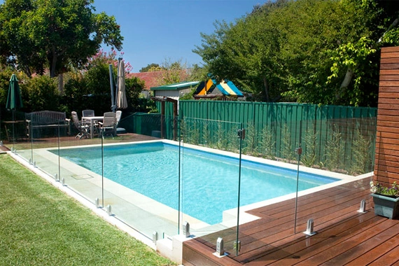 A swimming pool with a wooden deck and glass fencing.