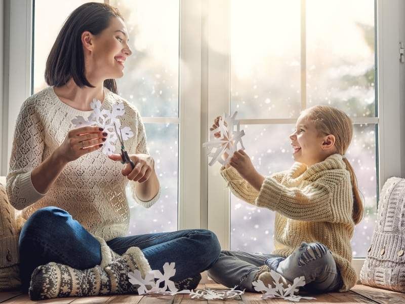 A mother and daughter enjoying a snowy home day by playing with snowflakes in front of a window.