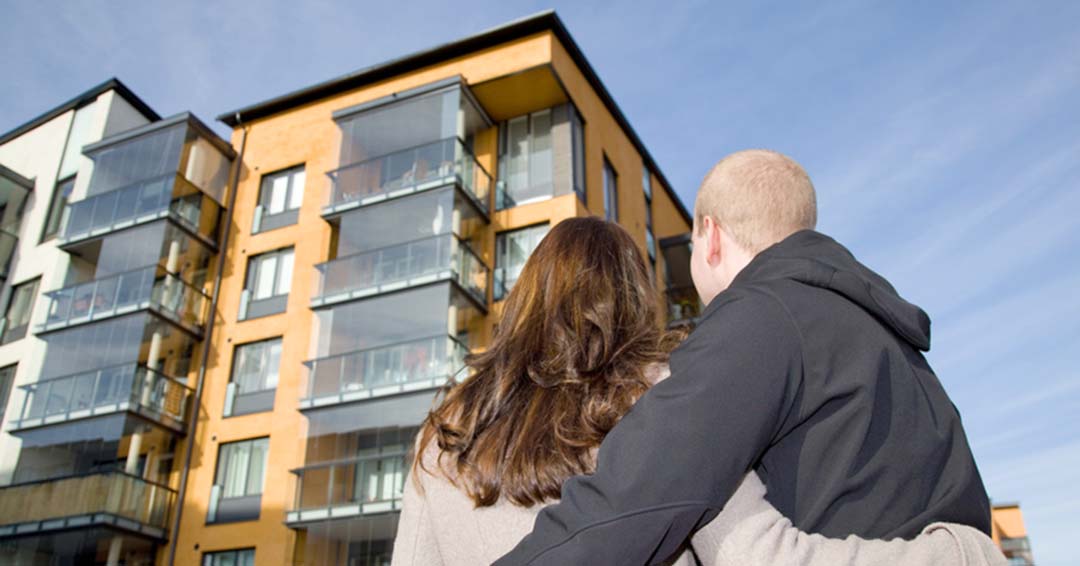 A couple hugging in front of an apartment building while considering buying a condo.